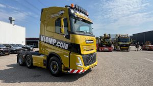 Zijkant van een gele Volvo vrachtwagen van klomp.com met opvallende zwarte en witte livery, geparkeerd op een industrieel terrein, met een andere vrachtwagen op de achtergrond.