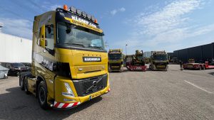 Voorkant van een gele Volvo vrachtwagen van klomp.com met opvallende zwarte en witte livery, geparkeerd op een industrieel terrein, met een andere vrachtwagen op de achtergrond.