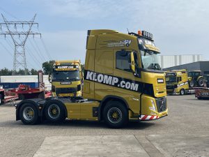 Zijkant van een gele Volvo vrachtwagen van klomp.com met opvallende zwarte en witte livery, geparkeerd op een industrieel terrein, met een andere vrachtwagen op de achtergrond.